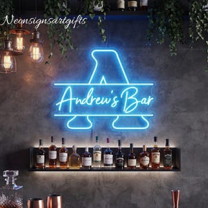 May include: A bright blue neon sign featuring a large letter A with a horizontal line through the center and the text Andrew's Bar written in a cursive script. The sign is mounted on a dark wall above a shelf holding various glass liquor bottles.