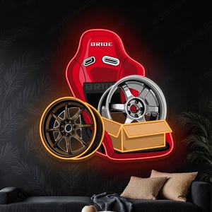 Pode incluir: Uma placa de néon vermelha com um assento de corrida com a palavra "BRIDE" e duas rodas de carro. Uma roda é castanha com contorno laranja, a outra é prateada. A placa está sobre um fundo preto.