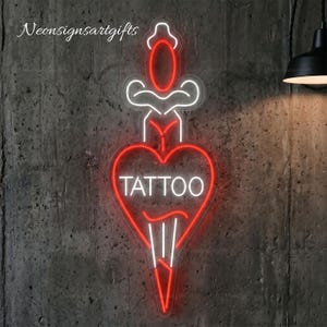 May include: A neon sign featuring a red and white heart shape pierced by a dagger, with the word TATTOO written in white capital letters inside the heart. The design includes a red oval at the top and white outlines forming the dagger and heart.