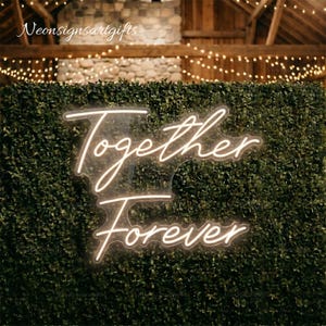 May include: A warm white neon sign featuring the script text Together Forever mounted on a clear acrylic backing. The sign is displayed against a dense green boxwood hedge wall, creating a bright contrast with the illuminated lettering.