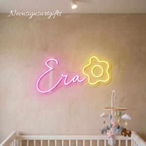 May include: A neon sign featuring the word Era in pink cursive script next to a yellow neon flower outline. The sign is mounted on a textured wall above a white crib with a hanging mobile featuring various shapes including a moon, stars, and animals.