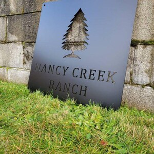 May include: Black metal sign with a cut-out pine tree design and the words "NANCY CREEK RANCH". The sign is square and mounted on a grassy area. The sign is leaning against a stone wall.