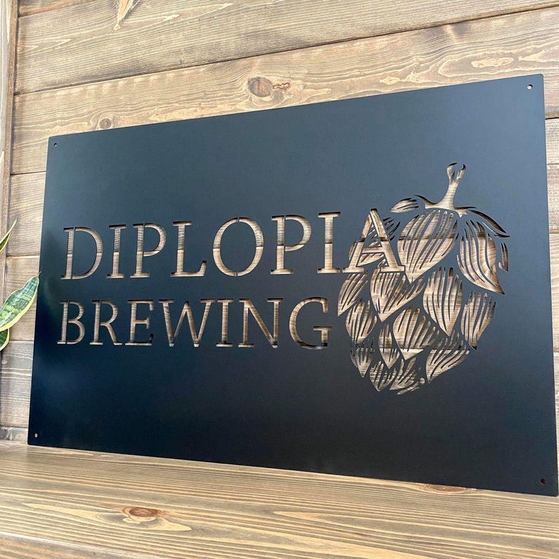 May include: Black metal sign with the words "DIPLOLIA BREWING" cut out, along with a hop flower design. The sign is mounted on a wooden surface and is suitable for a brewery or home bar.
