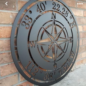 May include: A black metal compass rose wall art piece. The compass design includes directional indicators and coordinates. The piece is circular and mounted on a brick wall. The compass rose is cut out of the metal.