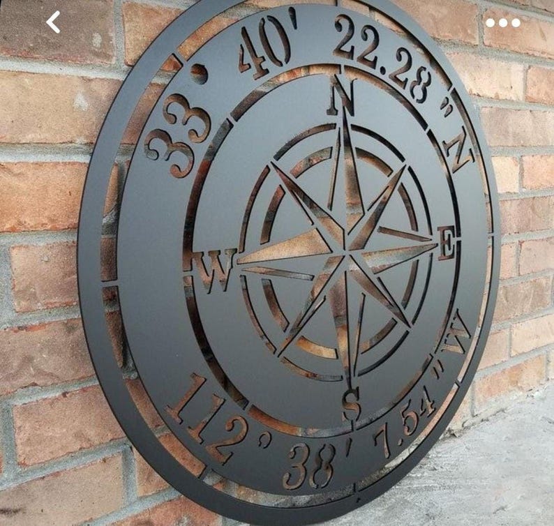 May include: A black metal compass rose wall art piece. The compass design includes directional indicators and coordinates. The piece is circular and mounted on a brick wall. The compass rose is cut out of the metal.