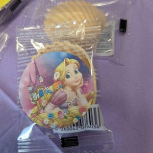 Peut inclure: Un biscuit emballé avec une image de Raiponce. Le biscuit est dans un emballage plastique transparent, sur un fond violet clair. L'image de Raiponce est sur un biscuit rond avec un château en arrière-plan.