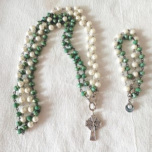 May include: A necklace and bracelet set featuring white faux pearls and green faceted beads. The necklace has a silver Celtic cross pendant. The bracelet has a small shell charm. The jewelry is displayed on a white surface.
