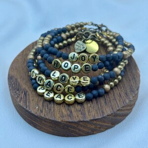 Pode incluir: Um conjunto de pulseiras de contas pretas e douradas. As pulseiras soletram as palavras "HOPE", "LOVE", "GRACE", "PEACE" e "JOY" com contas de letras douradas. Um pequeno pingente de coração dourado diz "Made with Love".
