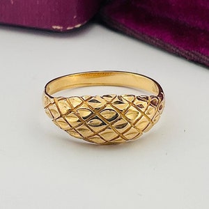 May include: A gold ring with a textured, diamond-shaped pattern on the top. The ring is sitting on a white surface next to two purple velvet boxes.