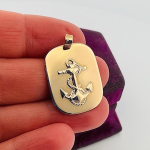 May include: A gold-colored rectangular pendant featuring a detailed anchor design. The anchor is centered on the pendant, with a rope wrapped around it. The pendant has a polished finish and a small loop at the top for a chain.
