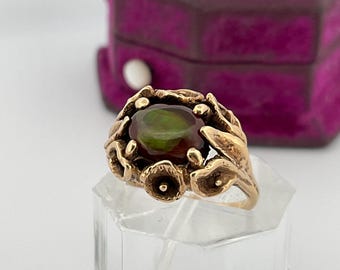 May include: A gold-toned ring featuring an oval-shaped, dark green gemstone. The ring's setting has a floral design with intricate details. The ring is displayed on a clear stand, with a purple velvet box in the background.