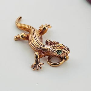 May include: A gold-toned lizard charm with green gemstone eyes. The lizard is detailed with textured scales and has a ring for attaching to a chain.