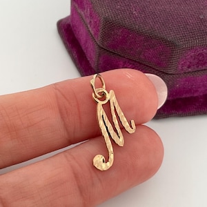 Può includere: Un ciondolo color oro a forma di lettera "M". Il ciondolo ha una superficie strutturata e un piccolo anello nella parte superiore per una catena. Lo sfondo include una scatola di velluto viola.