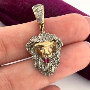 May include: A gold-toned lion pendant with a diamond-encrusted mane and bale. The lion's face is gold with ruby eyes and a ruby mouth. The pendant is held against a purple velvet background.