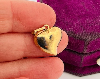 May include: A gold heart-shaped pendant charm. The charm is smooth and shiny, with a slight curve to the heart shape. The pendant is held in a hand, with a purple velvet box in the background.