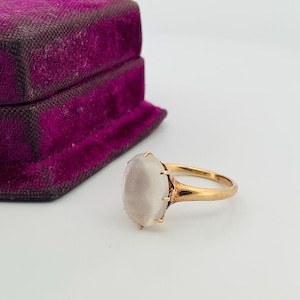 Pu&ograve; includere: Un anello d'oro con una pietra preziosa ovale bianco latte. L'anello &egrave; incastonato in un design classico, accanto a un portagioie vintage in velluto viola. La montatura e la fascia dell'anello sembrano essere in oro.