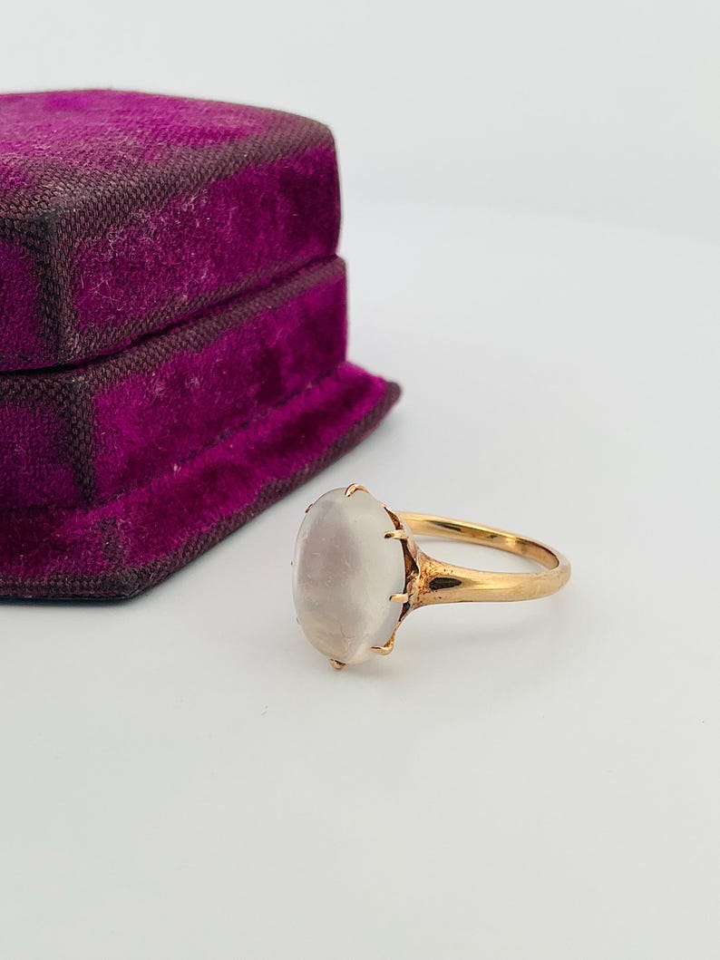 Pu&ograve; includere: Un anello d'oro con una pietra preziosa ovale bianco latte. L'anello &egrave; incastonato in un design classico, accanto a un portagioie vintage in velluto viola. La montatura e la fascia dell'anello sembrano essere in oro.