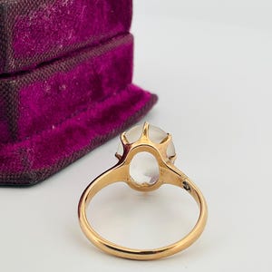 Pu&ograve; includere: Un anello d'oro con una grande gemma ovale e trasparente. L'anello &egrave; incastonato in uno stile classico con una fascia semplice. Un portagioie vintage in velluto viola &egrave; sullo sfondo.