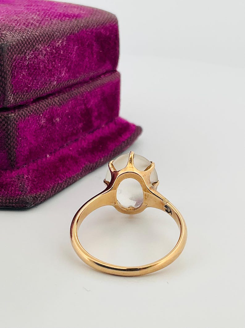 Pu&ograve; includere: Un anello d'oro con una grande gemma ovale e trasparente. L'anello &egrave; incastonato in uno stile classico con una fascia semplice. Un portagioie vintage in velluto viola &egrave; sullo sfondo.
