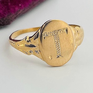 May include: A gold signet ring with an oval face bearing the letter "T". The ring has a detailed design on the band and face. The ring is displayed on a white surface.