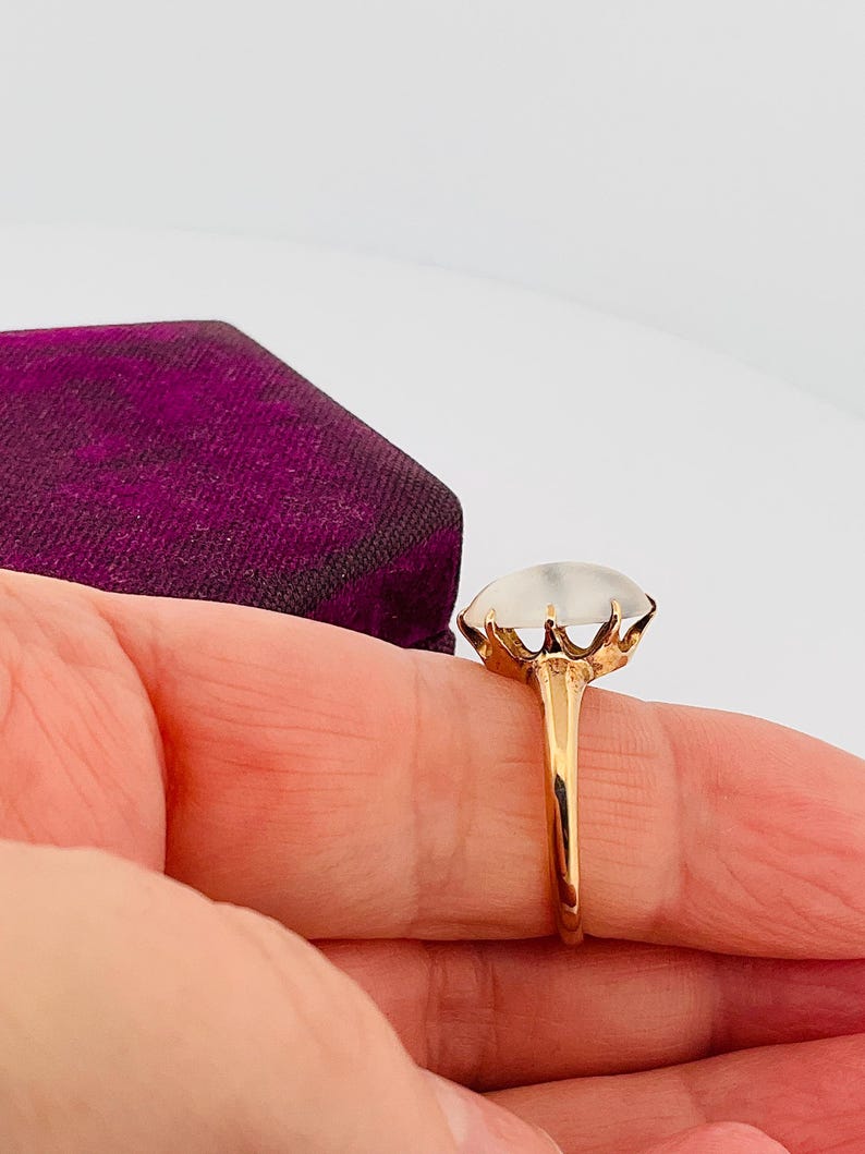Pu&ograve; includere: Un anello d'oro con una grande gemma ovale e traslucida. La gemma &egrave; incastonata in un'incastonatura a forma di corona e la fascia &egrave; in oro lucido. L'anello &egrave; tenuto contro uno sfondo di velluto viola.