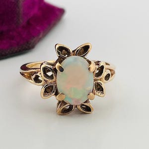 May include: A gold ring with an oval opal gemstone, set within a floral frame. The ring's band is gold, and the opal displays iridescent colours. The ring is on a white surface, with a purple velvet box in the background.