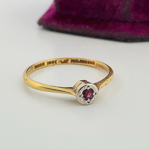 May include: A slender gold ring featuring a small, round, red gemstone in a silver-toned setting. The ring's band is thin and textured. The ring is displayed against a white backdrop.
