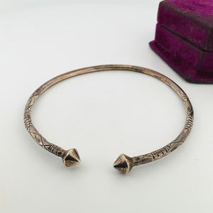 Peut inclure: Bracelet manchette ouvert en argent avec détails géométriques et texturés. Le bracelet a des embouts pointus et un motif gravé le long du bracelet. Une boîte à bijoux en velours violet est en arrière-plan.