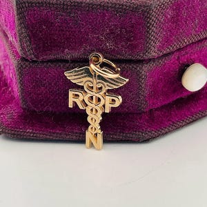 Op de afbeelding: Een goudkleurige RN-bedel met het Caduceus-symbool met vleugels en de letters R, N en P. De bedel wordt gepresenteerd tegen een donkerpaarse fluwelen achtergrond.