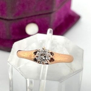 May include: A gold-coloured ring featuring a clear, round gemstone. The ring is displayed on a clear, angled stand. The ring's band has a simple, smooth design. The gemstone is the focal point, reflecting light.