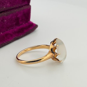 Pu&ograve; includere: Un anello d'oro con una grande gemma ovale bianco latte. La fascia e l'incastonatura dell'anello sono di un caldo color oro. La gemma &egrave; tenuta in posizione da sei griffe. Sullo sfondo, un portagioie di velluto viola.