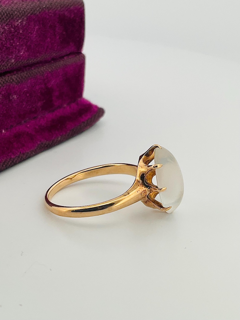 Pu&ograve; includere: Un anello d'oro con una grande gemma ovale bianco latte. La fascia e l'incastonatura dell'anello sono di un caldo color oro. La gemma &egrave; tenuta in posizione da sei griffe. Sullo sfondo, un portagioie di velluto viola.