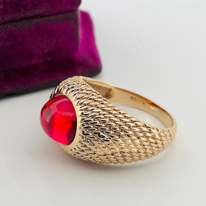Peut inclure: Une bague en or avec une bande texturée et une grosse pierre rouge ovale. La bague est posée sur un fond blanc avec un aperçu d'un écrin en velours violet.