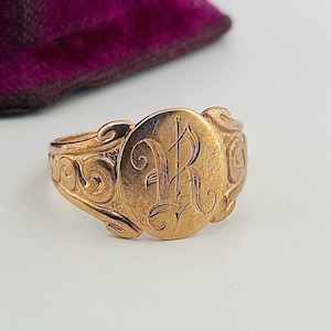 Peut inclure: Une bague chevalière dorée présentant une face ovale avec une lettre R gravée en écriture cursive. L'anneau affiche des motifs de volutes complexes sur les côtés, s'étendant depuis le sertissage ovale central. La surface métallique est polie.