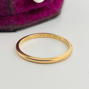 May include: A thin gold band ring with a smooth, polished finish. The interior of the band is engraved with the text BIRKS ELLIS 18K along with two small circular maker marks. The ring is positioned in front of a purple velvet jewelry box.