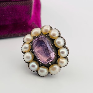 May include: An antique ring featuring a large, faceted, oval-cut amethyst gemstone at the center, surrounded by a halo of creamy white pearls. The ring has a silver-toned metal setting and is displayed on a white surface.