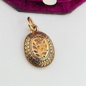 May include: An oval gold-coloured pendant featuring a floral design. The pendant has an engraved floral pattern in the centre, framed by a decorative border. It is suspended from a small loop.