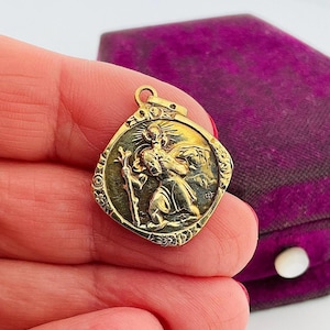 Könnte beinhalten: Ein goldfarbener Anhänger mit einem detaillierten Relief einer religiösen Figur, die ein Kind trägt. Der Anhänger hat ein kreisförmiges Design mit aufwendigen Details an den Rändern. Der Hintergrund umfasst eine lila Samtbox.