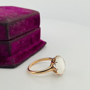 Pu&ograve; includere: Un anello d'oro con una grande pietra preziosa ovale bianco latte. L'anello &egrave; incastonato in uno stile classico con una fascia delicata. Un portagioie vintage in velluto viola &egrave; sullo sfondo.