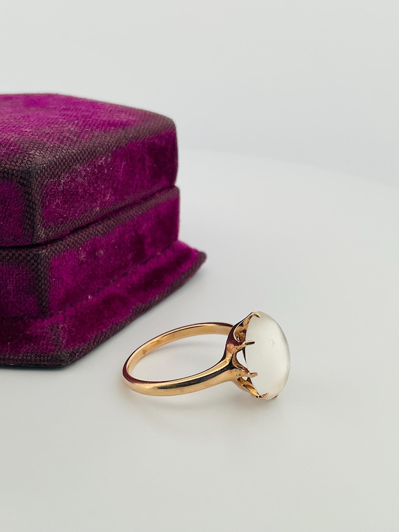 Pu&ograve; includere: Un anello d'oro con una grande pietra preziosa ovale bianco latte. L'anello &egrave; incastonato in uno stile classico con una fascia delicata. Un portagioie vintage in velluto viola &egrave; sullo sfondo.