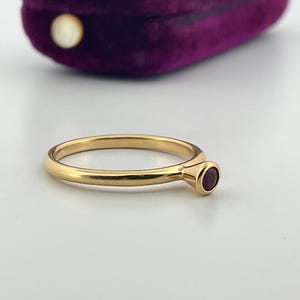 May include: A gold ring featuring a small, round, red gemstone. The ring is set against a white surface, with a purple velvet jewellery box in the background.