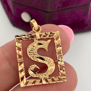 Peut inclure: Un pendentif en or présentant un cadre rectangulaire avec un motif texturé à rayures diagonales. Au centre du cadre se trouve une grande lettre S texturée. Le pendentif comprend une petite bélière en or sur le dessus pour une chaîne.