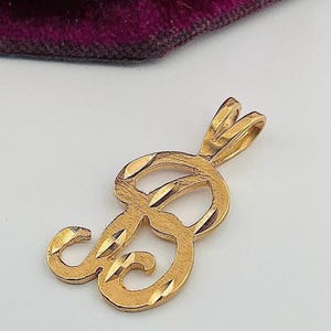 Peut inclure: Un pendentif doré en forme de lettre "B". Le pendentif présente un motif texturé taillé en diamant sur toute sa surface. La bélière est une simple boucle pour l'attacher à une chaîne. Le fond est de couleur claire avec une touche de violet.