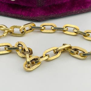 May include: A close-up shot of a gold chain necklace with oval links. The chain is a bright gold colour and appears to be made of metal. A portion of a purple velvet hat with a white button is visible in the background.