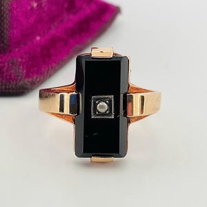 Könnte beinhalten: Goldfarbener Ring mit einem rechteckigen schwarzen Onyxstein und einem kleinen, quadratischen, silberfarbenen Akzent. Der Ring hat ein klassisches Design mit kreuzartiger Struktur, vor weißem Hintergrund.
