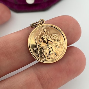 May include: A gold-coloured circular pendant featuring a detailed relief of a saint with a staff. It has a small loop for a chain. The background is a blurred purple velvet box.