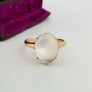Pu&ograve; includere: Un anello d'oro con una grande gemma ovale e traslucida. La fascia dell'anello &egrave; di un caldo colore oro e la gemma &egrave; tenuta in posizione da piccole griffe. L'anello &egrave; posto su uno sfondo bianco, con un portagioie di velluto viola sullo sfondo.