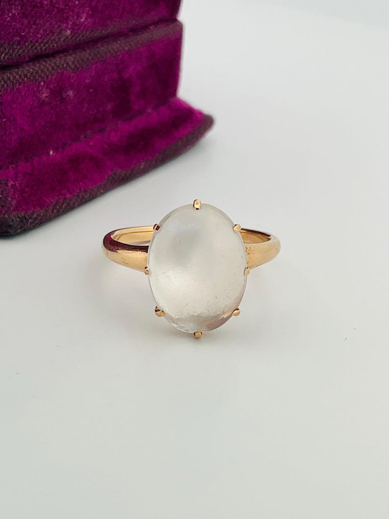 Pu&ograve; includere: Un anello d'oro con una grande gemma ovale e traslucida. La fascia dell'anello &egrave; di un caldo colore oro e la gemma &egrave; tenuta in posizione da piccole griffe. L'anello &egrave; posto su uno sfondo bianco, con un portagioie di velluto viola sullo sfondo.