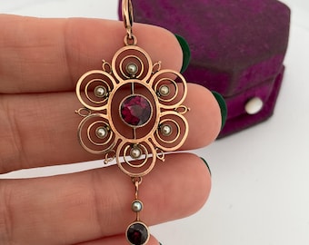 May include: A gold-toned pendant with a floral design, featuring a large, round, deep red gemstone at the center. The pendant has a series of concentric circles and small pearls. A smaller red gemstone hangs below.