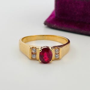 May include: A gold ring with a large oval ruby gemstone in the center, flanked by two smaller clear gemstones on each side. The ring has a simple, classic design and is set against a white background.